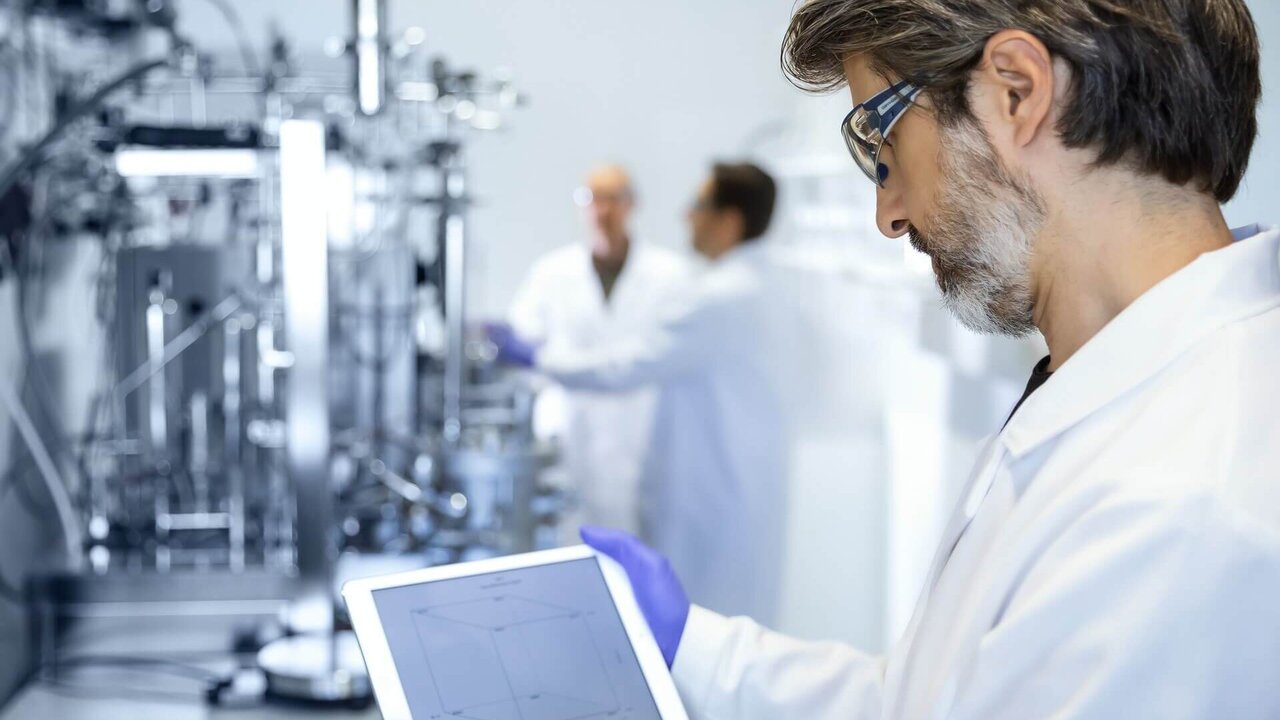 Scientist in a VALIDOGEN lab coat reviewing bioprocess data on a tablet in front of a stainless steel fermentation system, with colleagues optimizing fermentation parameters in the background